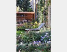 Ein gepflegter Garten mit blühenden Blumen in verschiedenen Farben, darunter violette und rote Blumen, sowie eine Liege im Vordergrund. Im Hintergrund ist eine mit Efeu bewachsene Backsteinwand und ein Holzunterstand mit Sitzmöbeln und Holzvorrat zu sehen.