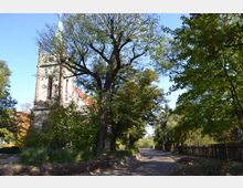 Ansicht einer kleinen Kirche mit spitzem Turm, umgeben von Bäumen. Im Vordergrund ist ein gepflasterter Weg zu sehen, der an der Kirche vorbeiführt, mit Zäunen und Vegetation auf beiden Seiten.