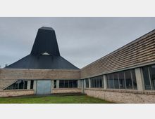 Ein modernes Gebäude mit einer dunklen, pyramidenartigen Dachstruktur und Wänden aus horizontal angeordneten Ziegelsteinen. Große Fensterfronten und ein Eingang aus Metall prägen die Fassade, umgeben von einer Wiese unter einem bedeckten Himmel.