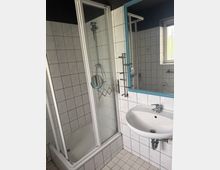Kleines, gefliestes Badezimmer mit Duschwanne und Glas-Schiebetür sowie einem Waschbecken neben einem großen Spiegel mit blauem Rahmen. Rechts sorgt ein Fenster für Tageslicht; die Wände und der Boden sind mit weißen Fliesen im Rastermuster versehen.