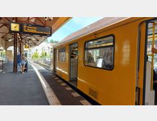 Gelber U-Bahn-Zug an einem überdachten Bahnsteig mit sichtbaren Gleisen, einem Zugzielanzeiger und einem Uhrzeitschild. Im Hintergrund sind Wohngebäude und Bäume zu erkennen, während Personen auf dem Bahnsteig stehen oder einsteigen.