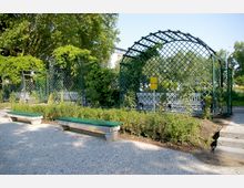 Öffentlicher Park mit einem gepflasterten Weg, zwei grünen Sitzbänken und einer rankenbewachsenen Metallpergola im Hintergrund. Die Umgebung ist von Büschen, Bäumen und einem offenen, sonnigen Himmel geprägt.
