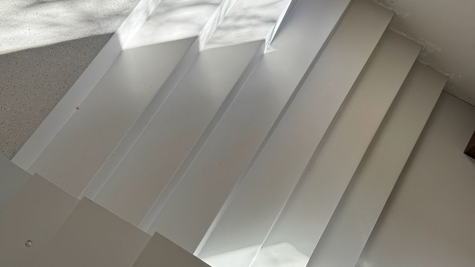 White indoor staircase with wide, rectangular steps leading down to a light gray speckled floor. Sunlight from a nearby opening casts sharp geometric shadows across the steps and adjacent wall.