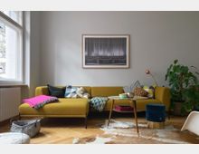 Wohnzimmer mit gelbem Ecksofa, dekoriert mit bunten Kissen und einer pinken Decke. Ein runder Couchtisch steht auf einem Kuhfell-Teppich, daneben ein blauer Hocker und eine Kupfer-Stehlampe; an der Wand hängt ein gerahmtes Bild mit Theatermotiv, im Hintergrund eine große Zimmerpflanze.