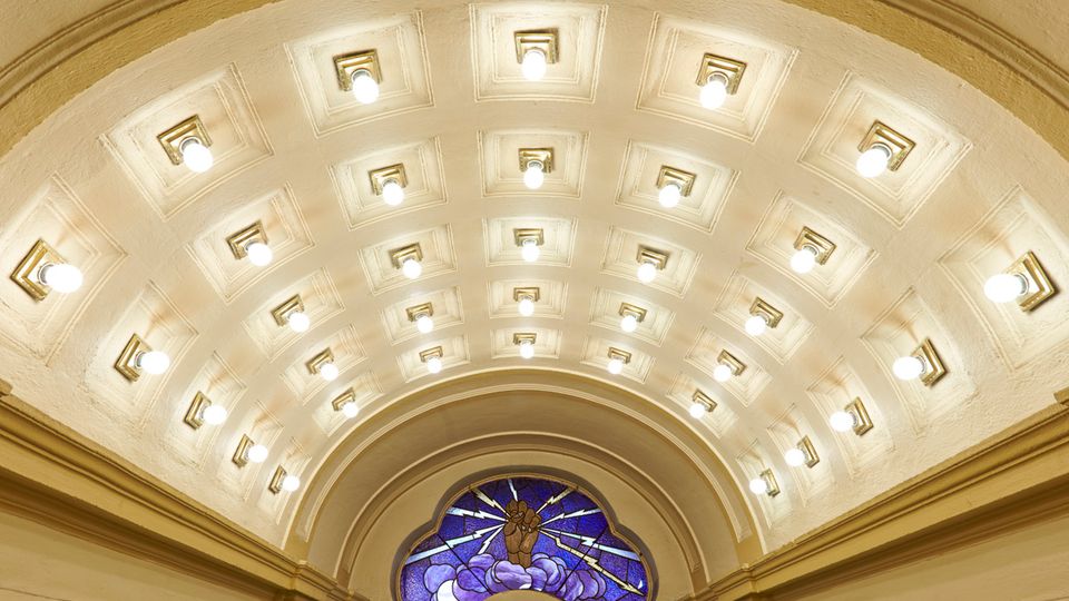 Gewölbedecke eines historischen Innenraums mit zahlreichen eingelassenen Lichtern in symmetrischer Anordnung. Unterhalb der Decke befindet sich ein buntes Glasfenster mit einer Darstellung einer erhobenen Hand und strahlenförmigen Mustern.