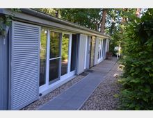 Langes, einstöckiges Gebäude mit weißen Schiebeläden und großen Fenstern, umgeben von Bäumen und Hecken. Vor dem Gebäude befindet sich ein schmaler Gehweg aus Pflastersteinen und Kies.