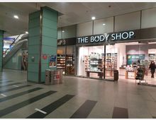 Ein Geschäftszentrum mit einer Filiale von „The Body Shop“. Die Ladenfront besteht aus Glas und zeigt helle Regale mit Kosmetikprodukten; links im Bild eine Rolltreppe und ein Poster an einer türkisfarbenen Säule.