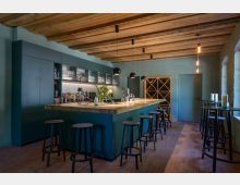 Eine moderne Bar mit einer langen, grünen Theke aus Holz, hohen schwarzen Barhockern und Hängelampen an einer Holzdecke. Im Hintergrund sind Regale mit Gläsern, Flaschen und ein Weinschrank zu sehen, der Raum hat Ziegelboden und eine rustikale Atmosphäre.