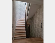 Treppe in einem Wohngebäude mit Holzstufen und Metallgeländer. Die Wände sind mit einer strukturierten Tapete in Grautönen gestaltet, und der Boden ist ebenfalls mit Holz verkleidet.
