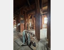 Historischer Innenraum einer Mühle mit Holzbalken, Holzboden und Müllerwerkzeug wie einer Waage im Vordergrund. Im Hintergrund sind Getreidesäcke, Holzkonstruktionen und ein Fenster mit Tageslicht sichtbar.