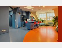 Ein modern gestalteter Aufenthaltsraum mit farbenfrohem Design, bestehend aus einer blauen Theke mit roten Barhockern, mehreren Sitzgelegenheiten, großen Fenstern mit Rollläden und zahlreichen Grünpflanzen. Der Boden ist in Blau und Orange gehalten, während die Wände überwiegend in Blau gestrichen sind.