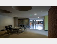 Innenraum eines modernen Gebäudes mit minimalistischer Einrichtung. Im Vordergrund steht ein schwarzer Tisch mit sechs Stühlen, im Hintergrund ist ein offener Bereich mit poliertem Boden, Sichtbetonpfeilern sowie einer Wand aus grauen Platten und einer roten Ziegelwand sichtbar.