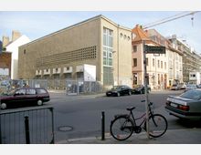 Ecke einer städtischen Straße mit einem grob verputzten Gebäude moderner Bauweise links, das rechteckige Lüftungsgitter und schmale Fenster hat. Rechts daneben befinden sich Wohnhäuser mit klassischen Fassaden und Baugerüste, im Vordergrund stehen geparkte Autos und ein Fahrrad an einem Straßenschild.