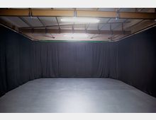 Ein leerer Raum mit schwarzem Vorhang an drei Wänden, grauem Boden und sichtbaren Metallträgern an der Decke, beleuchtet von grellem Deckenlicht.