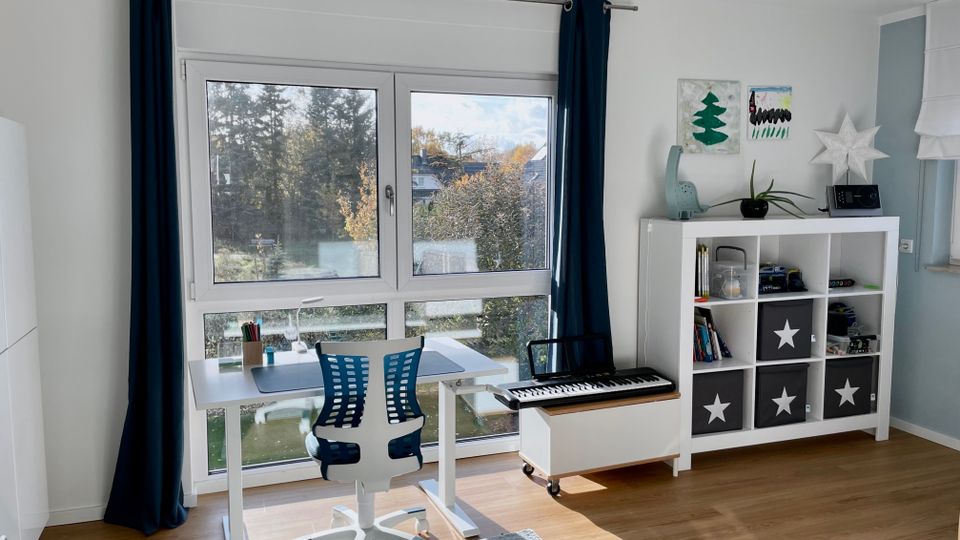 Heller Raum mit großer Fensterfront und dunklen Vorhängen, davor ein höhenverstellbarer Schreibtisch mit Bürostuhl. Rechts stehen ein weißes Regal mit Aufbewahrungsboxen und Dekoration sowie ein niedriges Möbel mit Keyboard; Holzboden und Blick ins Grüne.