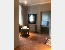 Ein rustikaler Innenraum mit terrakottafarbenem Fliesenboden, grauen Wänden und einem runden Tisch, der mit einer karierten Tischdecke bedeckt ist. Im Raum hängen Vorhänge vor einem Fenster, an der Wand befindet sich ein moderner Wandspiegel, und im Hintergrund ist eine Türöffnung zu einem anderen Raum sichtbar.