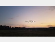 Ein kleines Flugzeug im Landeanflug bei Dämmerung, mit eingeschaltetem Landescheinwerfer und einem Wald im Hintergrund. Der Himmel ist in orange- und violettfarbenen Tönen gefärbt.