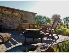 Steinterrasse mit Sitzmöbeln aus Holz und Rattan vor einer rustikalen Natursteinmauer. Im Vordergrund steht eine runde, erhöhte Feuerschale, im Hintergrund sind Bäume und eine sonnige Wiese sichtbar.