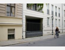 Mehrfamilienhaus in einer Stadtstraße mit hellem Putz und schmalen Fenstern; eine metalschwarze Einfahrt mit Tor führt zu einem Innenhof. Im Vordergrund fährt eine Person auf einem Fahrrad auf der gepflasterten Straße.