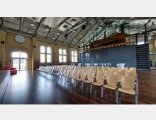 Großer Innenraum mit einer modernen, industriellen Architektur, gekennzeichnet durch freiliegende Stahlträger und eine Decke mit Beleuchtungselementen. Der Raum hat helle Backsteinwände, große Fensterbögen, eine Bühne links und zahlreiche Holzstühle in Reihen auf einem dunkelbraunen Holzboden.
