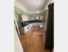 Geräumige Küche mit Fischgrät-Holzboden, weißen Hängeschränken und einer Arbeitsfläche aus dunklem Material. Links befinden sich eine Waschmaschine und ein Trockner, rechts ein großer Kühlschrank; in der Mitte steht ein Holztisch auf einem Teppich.