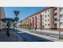 Straßenszene mit mehrstöckigen Wohngebäuden, die beige Wände und rote Balkone haben, sowie einem Bürgersteig mit einer Bushaltestelle mit überdachtem Wartebereich im Vordergrund. Am Straßenrand stehen Bäume, und ein Straßenschild zeigt „Brandenburger Straße“ an.