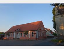 Ein einstöckiges Gebäude mit orangefarbener Fassade, weißen Fensterrahmen und einem roten Satteldach steht an einer gepflasterten Straße. Im Vordergrund sind Laternen und kleine Bäume zu sehen, rechts grenzt ein weiteres größeres Gebäude an.