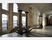 Großer Innenraum mit klassischer Architektur, bestehend aus hohen Fenstern mit Buntglas, verzierten Säulen, geschwungenem schmiedeeisernem Treppengeländer und gewölbten Decken. Der Raum ist in hellen Tönen gehalten und von natürlichem Licht durchflutet.