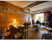 Gemütlicher Café- oder Barinnenraum mit Holzvertäfelung, gemischten Tischen und Stühlen sowie einem großen Wandbanner „LIEBE, SCHNAPS & REVOLUTION“. Sichtbare Lüftungsrohre an der Decke und ein großes Fenster zur Straße sorgen für industriellen Charakter und Tageslicht.