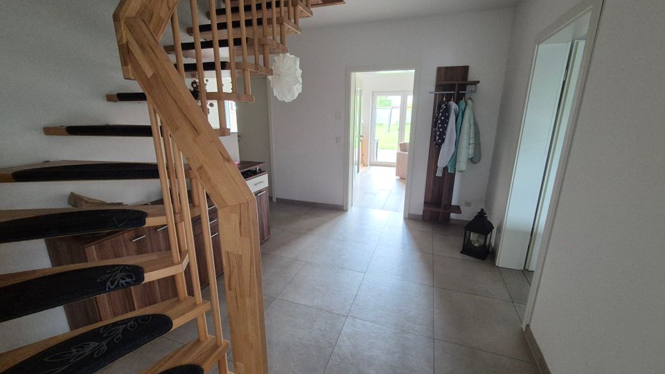 Heller Eingangsbereich eines Wohnhauses mit großer, grauer Fliesenbodenfläche und einer offenen Holztreppe mit Geländer. Rechts stehen eine Garderobe und ein großer Spiegel; durch eine Türöffnung ist ein weiterer Raum mit Blick nach draußen zu sehen.