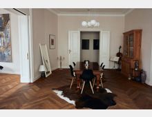 Esszimmer in einem Altbau mit Parkettboden, einem dunklen Holztisch und sechs schwarzen Stühlen auf einem Fellteppich. Im Hintergrund stehen ein Holzvitrinenschrank, ein Cello mit Notenständer sowie dekorative Elemente wie ein Spiegel und Kerzenhalter.