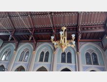 Gotischer Innenraum einer Kirche mit spitzbogigen Fenstern, verzierten Glasmalereien und einem reich dekorierten Holzdach. Im Vordergrund hängt ein goldener Kronleuchter mit weißen Kerzenlampen.