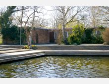 Backsteingebäude mit flachem Dach, umgeben von Bäumen und Sträuchern. Im Vordergrund befinden sich Holzstege und ein Gewässer mit ruhiger Wasseroberfläche.