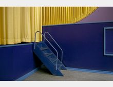 Eine kleine dunkelblaue Treppe mit Geländern führt zu einem orange-gelben Vorhang vor einer dunkelblauen Wand mit minimalistischer Gestaltung. Der Boden besteht aus einem grauen Teppichbelag.