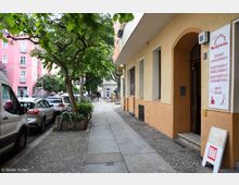 Straßenszene in einer Wohngegend mit Bürgersteig, bepflanzten Bäumen, mehreren geparkten Autos und einem Gebäude mit hellgelber Fassade. Rechts sind ein Ladeneingang und ein Schild mit Werbung für Backwaren zu sehen.