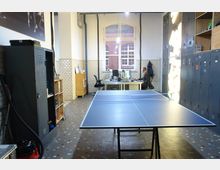 Büro mit industriellem Flair, ausgestattet mit einem Tischtennistisch im Vordergrund, Schließfächern an den Wänden sowie einem Arbeitsplatz mit Schreibtisch und Computer vor einem großen Fenster mit Blick auf ein Backsteingebäude. Die Wände sind mit weißen Fliesen verkleidet, und der Boden besteht aus farbigen Mosaikfliesen.