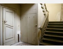 Eingangsbereich eines alten Gebäudes mit einer hellen Holztür links, einer verschlossenen Holzverkleidung in der Mitte und einer abgenutzten Holztreppe mit einem verzierten Geländer rechts. Der Boden ist mit gemusterten Fliesen ausgelegt, und die Wände zeigen Gebrauchsspuren.