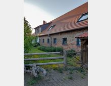 Ein Bauernhaus aus rotem Ziegelstein mit einem steilen roten Ziegeldach und mehreren Dachfenstern. Im Vordergrund befindet sich ein kleiner Garten, der durch einen einfachen Holzzaun begrenzt ist.