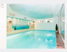 Innenpool in einem hellen Raum, mit Wänden, die eine mediterrane Landschaftsmalerei zeigen. Der Raum hat mehrere kleine Fenster mit Pflanzen und dekorativen Elementen, während der Boden um den Pool aus Marmor besteht.