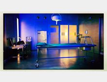 Ein steriler Raum mit kühler Beleuchtung, der wie eine Pathologie oder ein Labor wirkt. Zu sehen sind ein Metalltisch mit Rädern, eine Arbeitsstation mit Monitor und Lampe, sowie eine Tür mit strukturiertem Metall und Fenstern im Hintergrund.