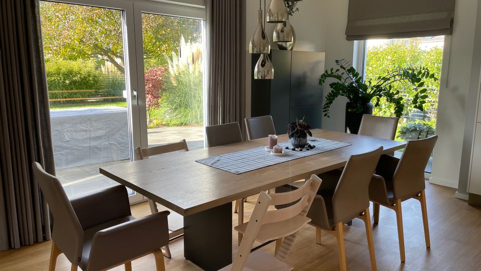 Esszimmer mit einem großen Holztisch und acht beigefarbenen Stühlen, beleuchtet durch eine Hängelampe mit mehreren silbernen Elementen. Große Fenster mit Blick auf einen Garten mit Bäumen, Sträuchern und Gräsern; dunkle Vorhänge und ein heller Holzfußboden ergänzen das moderne Design.