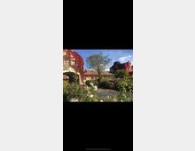 Historische Gebäude mit roten Kletterpflanzen an den Fassaden, umgeben von einem Garten mit bunten Blumen und einem gepflasterten Weg. Ein großer Baum erhebt sich in der Mitte des Gartens unter einem klaren blauen Himmel.