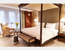 Hotelzimmer mit einem Himmelbett, Nachttischen mit Lampen, einem gepolsterten Hocker und einer Sitzecke mit Sessel und Beistelltisch. Große Fenster mit bodenlangen Vorhängen sorgen für Helligkeit, und eine Kommode aus Holz steht an der Wand.