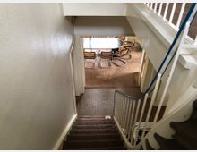 Treppenhaus in einem Wohngebäude mit braunem Teppichboden und weiß gestrichenen Wänden; im Hintergrund ein Raum mit einem ovalen Esstisch und mehreren Stühlen auf Teppichboden.