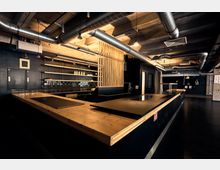 Industrieküche mit modernem Design, dominierend durch Holz- und Metallakzente. Sichtbare Lüftungsrohre an der Decke, offene Regale, eine Arbeitsfläche aus Holz und dunkle Fliesen an der Rückwand prägen den Raum.