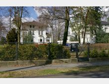 Weiße Villa im Stil der Gründerzeit, umgeben von Bäumen und einer gepflegten Gartenanlage, hinter einem Metallzaun mit Tor und Klingelanlage. Im Vordergrund eine Straße mit Gehweg, im Hintergrund ein weiterer Gebäudeteil.