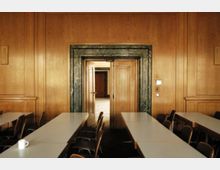 Ein Besprechungsraum mit Holzvertäfelung an den Wänden, langen weißen Tischen und Stühlen. Im Vordergrund führt eine Doppeltür mit grünem Steinumrahmungen in einen angrenzenden Raum.