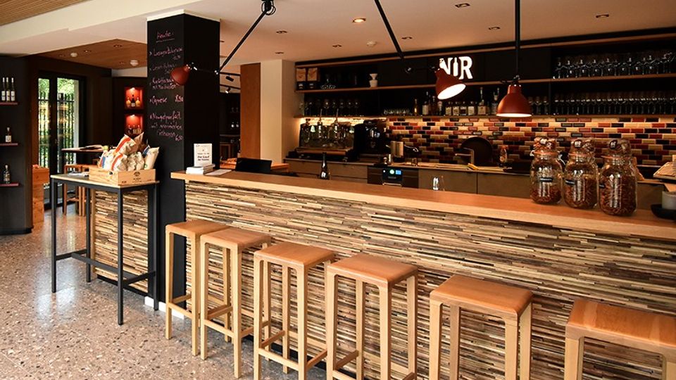 Moderner Café- und Barbereich mit langem Tresen in Holzoptik und hohen Barhockern davor. Im Hintergrund sind Regale mit Gläsern, eine Kaffee-Station, eine mehrfarbige Fliesenrückwand sowie Pendelleuchten und eine Tafelwand zu sehen.