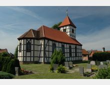 Fachwerkkirche mit roten Ziegeldächern und einem Glockenturm in einem ländlichen Umfeld, umgeben von einem gepflegten Friedhof mit Grabsteinen und blühenden Pflanzen. Im Hintergrund sind weitere Gebäude und ein blauer Himmel zu sehen.