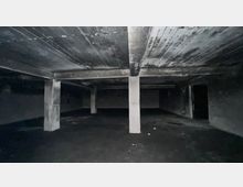 Ein leerer, dunkler Betonraum mit mehreren Sichtbetonpfeilern, einer niedrigen Decke, sowie kahlen Wänden. Der Boden und die Decke weisen Spuren von Feuchtigkeit und Verfall auf.